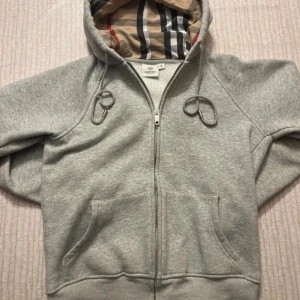 Grå Burberry hoodie med dragkedja - Snygg grå hoodie från Burberry med klassiskt rutigt foder i huvan och dragkedja framtill. Tröjan har snörning i huvan, känguruficka och är tillverkad i mjuk bomull. Perfekt för dig som vill ha en stilren och lyxig vardagslook. Storlek M men sitter mer som S-Xs
