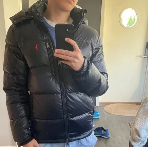 Svart dunjacka från Polo Ralph Lauren - Snygg glansig svart dunjacka från Polo Ralph Lauren med röd logga på bröstet. Jackan är i riktigt bra skick, bara en liten lagning på undersidan av luvan, så inget som syns vid användning. Tveka inte med frågor!