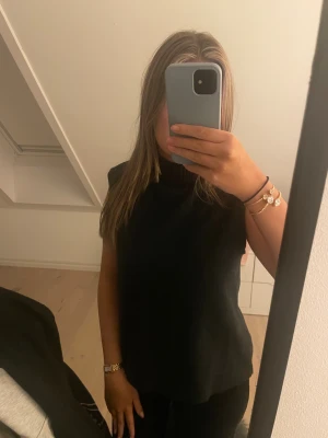 Svart polotopp i stickad material - Säljer en svart polotopp i stickad material med ärmlös design. Toppen har en hög krage och en loose passform som ger en chill och stilren vibe. Perfekt att matcha med jeans eller kjol för en enkel men snygg look.