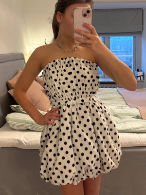 Prickig klänning - En kort axelbandslös klänning i vit med svarta prickar. Ballongformad kjol och rynkad överdel som är perfekt som temafester! Klänningen har markerad midja och är i ett lätt, mjukt tyg.