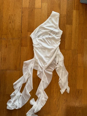 Vit asymmetrisk klänning från Bebe M - Supersnygg vit kortklänning från Bebe med ett axelband och glittrande Bebe-logga framtill. Klänningen har transparenta volangdetaljer i mesh som ger en unik look. Perfekt för dig som vill sticka ut med en edgy och trendig stil.