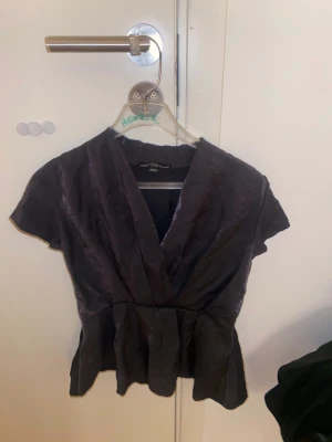 Svart omlottblus från XOXO Friends - Snygg svart omlottblus från XOXO Friends i glansigt tyg. Blusen har korta ärmar, markerad midja och lätt volang nertill. Perfekt för dig som gillar en stilren look med lite extra shine. Passar dig som vill ha något unikt och trendigt.