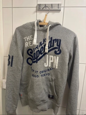 Superdry hoodie  - Grå hoodie från Superdry med stort blått och vitt tryck på framsidan, samt nummerdetalj på ärmen. Tröjan har huva med vita snören och en klassisk magficka. Mjuk insida och ribbade muddar vid ärmslut och nederkant. Aldrig använd 