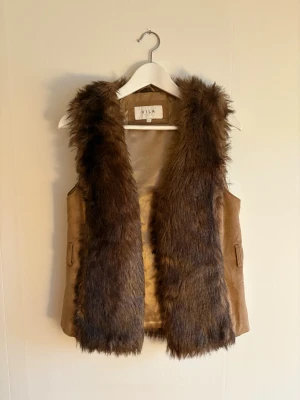 Brun pälsväst från VILA, strl M - Säljer en snygg brun pälsväst från VILA i storlek M. Västen har fluffig fuskpäls framtill och baktill, med sidor i mjuk mockaliknande material. Fodret är glansigt och beige. Saknar bältet.