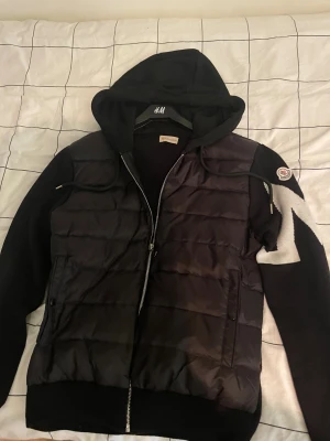 Svart Moncler pufferjacka med huva - Säljer en svart pufferjacka från Moncler med huva och dragkedja. Jackan har stickade ärmar med vit zigzag-mönster och Moncler-logga på ena ärmen. Kroppen är quiltad och vadderad, perfekt för kyligare dagar. Snygg och sportig design som sticker ut.