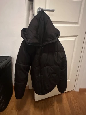 Svart pufferjacka med huva - Säljer en svart pufferjacka med stor huva och dragkedja framtill. Jackan är quiltad och har en loose passform, perfekt för kalla dagar. Fickor på sidorna och dragsko i huvan för extra komfort. Enkel och stilren design som funkar till det mesta.