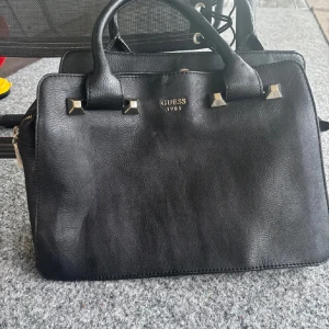 Svart handväska från Guess i skinn - Stilren svart handväska från Guess med guldfärgade metalldetaljer och logga framtill. Väskan har två handtag, flera fack och dragkedja i mitten. Tillverkad i skinn med en klassisk, fyrkantig form som passar till många outfits. Se slitningar på bilderna😁 Använd gärna köp nu❤️