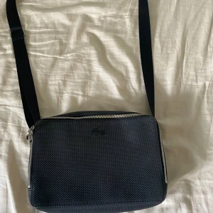 Svart axelväska från Lacoste - Snygg svart axelväska från Lacoste med diskret krokodillogga framtill. Väskan har en strukturerad yta, justerbar axelrem och dragkedja upptill. Invändigt finns ett grönt foder och praktiska fack, inklusive ett med dragkedja. Perfekt för att bära det viktigaste.