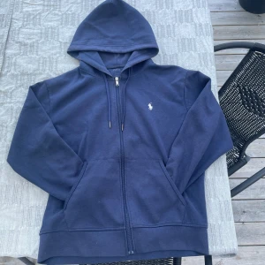 Marinblå hoodie från Polo Ralph Lauren - Marinblå hoodie från Polo Ralph Lauren med dragkedja, huva med snörning och klassisk vit broderad logga på bröstet. Tröjan har känguruficka framtill och är tillverkad i mjuk bomull. Perfekt för chill dagar och snygg streetstil.