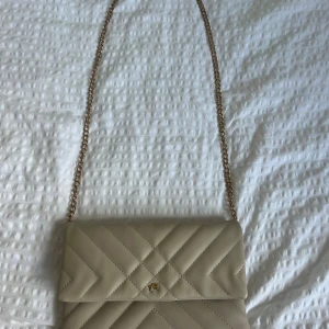 Beige axelväska med kedja - Snygg beige axelväska! Väskan har ett stilrent kuvertformat lock och liten gulddetalj framtill. Perfekt för dig som vill ha en trendig och enkel väska i skinnimitation. Dessvärre har den lite defekter på kedjan och en fläck inuti men jag skulle tro att den går att tvätta bort!