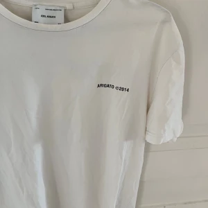 Vit t-shirt från Axel Arigato - Snygg vit t-shirt från Axel Arigato med svart tryck både fram och bak. Klassisk passform med rund hals och korta ärmar. Tillverkad i mjuk bomull, perfekt för en clean och stilren look. Trycket har texten 'ARIGATO ©2014' och adressdetaljer på ryggen.