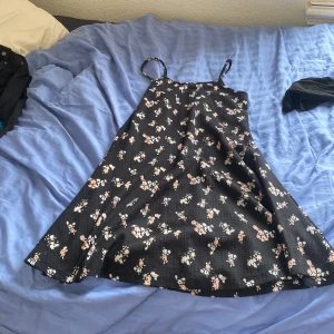 Svart blommig kortklänning 157 New - Supersöt svart kortklänning med tunna axelband och blommigt mönster i rosa och vitt från 157 New. Klänningen har en lätt utställd passform och är perfekt för varma dagar. Materialet känns mjukt och bekvämt mot huden.