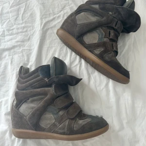 Isabel Marant sneakers brun/grå - Säljer ett par Isabel Marant sneakers i brunt och grått mocka med coola kardborreband. Skorna har en dold kilklack på ca 5 cm och gummisula, perfekt för dig som vill ha både stil och lite extra höjd. Ikonisk streetstyle-vibe och riktigt snygga detaljer. Självklart äkta! Hör gärna av er vid frågor 🙌🙌
