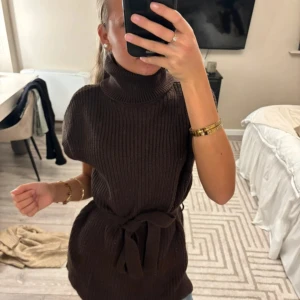 Brun stickad tröja med polokrage - Mysig brun stickad tröja med polokrage och knytskärp i midjan. Modellen är ärmlös och har en ribbad struktur som ger en cool och avslappnad vibe. Perfekt för lager-på-lager och passar snyggt till jeans eller tights.