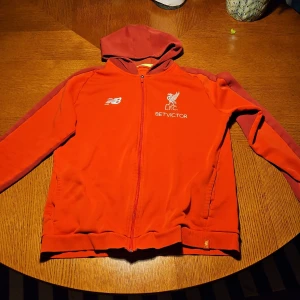 Liverpool hoodie från New Balance röd - Röd Liverpool FC hoodie från New Balance med broderad logga och BETVICTOR-tryck på bröstet. Tröjan har huva, dragkedja framtill och långa ärmar i en något mörkare röd nyans. Perfekt för fotbollsfans som vill visa sitt lag.