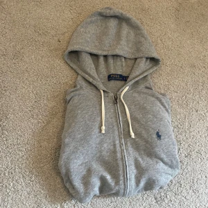 Grå hoodie från Polo Ralph Lauren - Säljer en grå hoodie från Polo Ralph Lauren med dragkedja och huva. Tröjan har vita snören, två fickor fram och den klassiska broderade loggan i mörkblått på bröstet. Perfekt för chill dagar och enkel att matcha med jeans eller joggers.