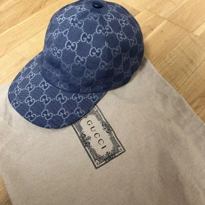 Blå Gucci keps med GG-mönster - Snygg blå keps från Gucci med ikoniskt GG-mönster över hela kepsen. Kepsen är tillverkad i bomull och har en klassisk böjd skärm samt en liten blå knapp på toppen. Insidan är vit och har Gucci-etikett. Perfekt för dig som vill ha en lyxig streetstyle-accessoar.