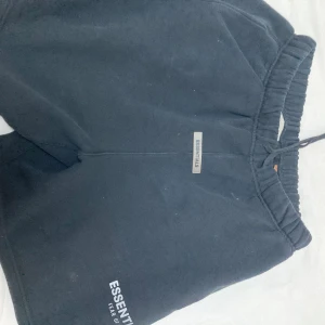 Svarta Essentials shorts Fear of God - Svarta shorts från Essentials Fear of God med broderad logga framtill. Modellen har elastisk midja med snörning och är tillverkad i mjuk bomull. Perfekta för chill eller träning, klassisk loose fit och stilren design.