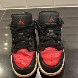 Nike Air Jordan 1 Low svart/röd 38 - Säljer ett par Nike Air Jordan 1 Low i klassisk svart och röd färg med vita detaljer. Skorna har snörning, rund tå och är tillverkade i skinn och syntet. Ikonisk Jumpman-logga på plösen och Air Jordan-märke på hälen. Perfekta för dig som gillar streetwear och sneakers med attityd.