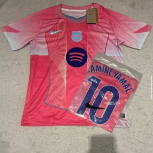 Barca rosa fotbollströja Nike Lamine Yamal - Säljer en FC Barcelona fotbollströja i rosa och ljusblått från Nike med Lamine Yamal och nummer 10 på ryggen. Tröjan har korta ärmar, tryckt klubbmärke och sponsorlogga på bröstet samt coola grafiska linjer framtill. Materialet är lätt och andas, perfekt för match eller träning.