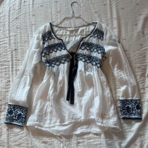 Vit blus med blå broderi Odd Molly - Säljer en vit blus från Odd Molly med blå broderade detaljer på bröstet och ärmsluten. Blusen har lång ärm, v-ringning och ett snyggt knytband framtill. 