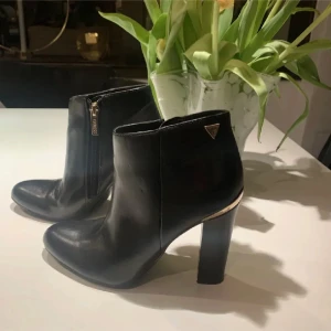 Svarta stövletter från Guess med gulddetalj - Snygga svarta stövletter från Guess i skinn med guldfärgade detaljer vid klacken och dragkedja på sidan. Skorna har rund tå och hög klack, samt en diskret Guess-logga på utsidan. Perfekta för dig som vill ha en stilren och trendig look.