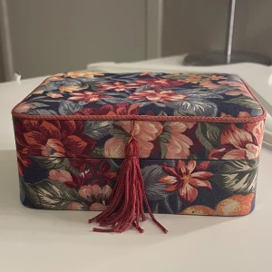 Blommig smyckesask med spegel - Superfin smyckesask med blommigt mönster. Asken har en dekorativ röd tofs framtill och en inbyggd spegel i locket. Perfekt för att förvara ringar, armband och örhängen.