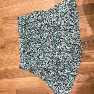 Blommig kortkjol från Zara - Supersöt blommig kortkjol från Zara i vitt, svart och turkost. Kjolen har volangig nederkant och ett lätt, flowy tyg som känns perfekt för varma dagar. Sitter snyggt i midjan och har ett feminint, lekfullt mönster.