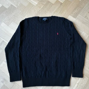 Navy blå kabelstickad tröja Polo Ralph Lauren - Kabelstickad svart tröja från Polo Ralph Lauren i 100% bomull. Klassisk rund halsringning, ribbade muddar och nederkant samt den ikoniska röda loggan broderad på bröstet. Perfekt för dig som gillar stilren och tidlös design. Det står Xl men det är en speciell storlek som är mer för barn och är ungefär som S i vuxen storlek
