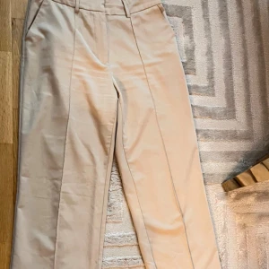 Beiga kostymbyxor från NA-KD - Stilrena beiga kostymbyxor från NA-KD med pressveck och hällor för bälte. Byxorna har rak passform och är tillverkade i ett mjukt bomullsmaterial. Perfekta för en clean och snygg look.