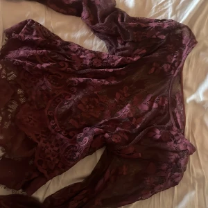 Vinröd blommig spetsblus - Säljer en vinröd blus i transparent spets med blommigt mönster. Blusen har lång ärm och en loose passform, perfekt för dig som gillar detaljer och romantisk stil. Materialet är mjukt och följsamt, med vacker spets och blommiga inslag.