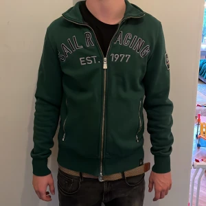 Grön ziptröja från Sail Racing  - Mörkgrön ziptröja från Sail Racing med broderad logga och text på bröstet. Tröjan har hel dragkedja, två fickor med dragkedja och patch på ena ärmen. Tillverkad i 80% bomull och 20% polyester. Perfekt för dig som gillar sportig och stilren look. Modellen är 178 cm lång och väger 63 kg. Det är bara att höra av sig vid frågor och funderingar 😀