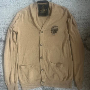 Beige kofta från Morris  - Snygg beige kofta från Morris  med broderad logga och text på bröstet. Koftan har v-ringning, knäppning framtill med fyra knappar och två fickor. Perfekt för lager-på-lager och stilrena outfits.