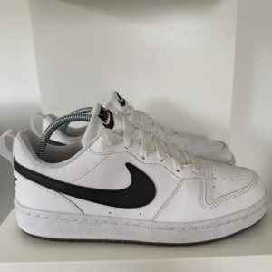 Nike Air Force 1 vita sneakers - Säljer ett par klassiska Nike Air Force 1 sneakers i vitt skinn med svart swoosh. Skorna har rund tå, snörning och platt sula. Perfekta för dig som gillar en clean och tidlös look. Passar till det mesta och är riktigt bekväma.
