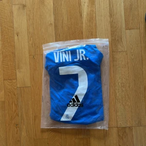 Real Madrid Vini Jr. blå fotbollströja - Blå Real Madrid fotbollströja från Adidas med Vini Jr. och nummer 7 på ryggen. Tröjan har vita detaljer, broderat klubbmärke, Adidas-logga och sponsortryck på bröstet. Tillverkad i lätt och ventilerande polyester med klassisk rund hals och korta ärmar.