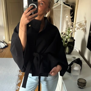 Svart cape poncho - Säljer en svart cape med breda ärmar och snygg omlottknäppning. Jackan har ett bälte i midjan som ger en schysst siluett och en guldig knapp vid halsen. Perfekt för dig som gillar stilrena och trendiga plagg.