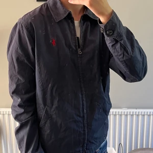  Polo Ralph Lauren jacka - Snygg mörkblå vindjacka från Polo Ralph Lauren med klassisk krage och röd broderad logga på bröstet. Jackan har dragkedja framtill, två snedställda fickor och långa ärmar med knapp vid ärmslut. Perfekt för dig som gillar stilren och tidlös design. Har tecken på användning 