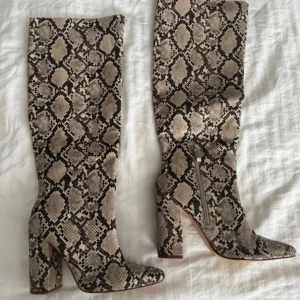 Höga boots med ormskinnsmönster - Säljer ett par höga boots med chunky klack och spetsig tå i beige och brunt ormskinnsmönster. Skorna har dragkedja på insidan och är tillverkade i syntetmaterial som imiterar ormskinn. Perfekta för dig som vill sticka ut med en edgy vibe.