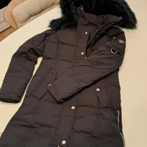 Svart lång Rockandblue dunjacka med päls - Säljer en svart lång dunjacka från Rockandblue med stor huva och fluffig päls. Jackan har dragkedja, tryckknappar, fickor och snygga detaljer på ärmen. Perfekt för kalla dagar när du vill hålla stilen och värmen.