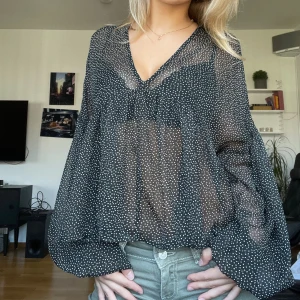 Svart prickig blus från NA-KD - Säljer en luftig svart blus från NA-KD med vita små prickar, v-ringning och volangdetaljer. Blusen är transparent med långa, vida ärmar och har en flowy passform som ger en cool och avslappnad vibe.