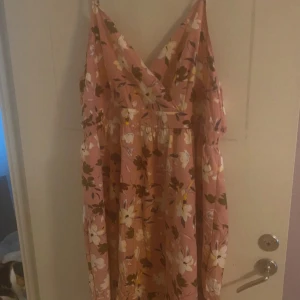 Blommig rosa långklänning - Söt långklänning i rosa med stora vita och gula blommor samt gröna blad. Klänningen har v-ringad omlott-topp och smala axelband. Perfekt för sommarens varma dagar och har en luftig, flowy passform.