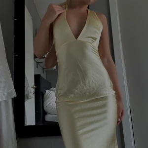 Ljusgul långklänning med djup v-ringning - Supersnygg långklänning i ljusgul satin. Klänningen har djup v-ringning, halterneck och öppen rygg. Sitter figurnära och har en elegant, slät finish som ger en lyxig känsla. Perfekt balklänning eller vid andra specifika tillfällen. Går lite över fötterna 