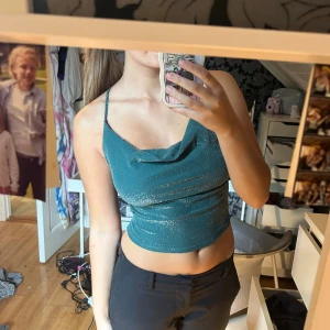 Glittrig grön croppad linnetopp - Säljer en glittrig grön croppad linnetopp med smala axelband och snygg draperad halsringning. Toppen har öppen rygg med snörning och knyt, vilket ger en cool och trendig look. Perfekt för dig som vill sticka ut med lite extra glitter.