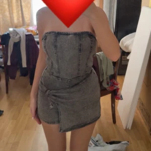 Grå tubklänning i jeanslook - Säljer en grå, tight tubklänning i jeanslook med asymmetrisk nederdel och markerade sömmar. Klänningen är kort, axelbandslös och har dragkedja bak. Perfekt för dig som gillar edgy och trendig stil.