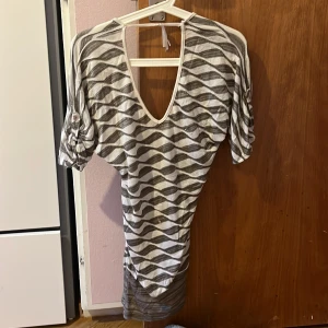 Randig vintage kortklänning med snörning - Säljer en kortklänning med grått och vitt vågigt randigt mönster. Klänningen har v-ringad hals, korta puffärmar och snygg snörning framtill. Materialet känns mjukt och lätt, perfekt för varmare dagar. Passformen är normal och klänningen har en cool, modern vibe.