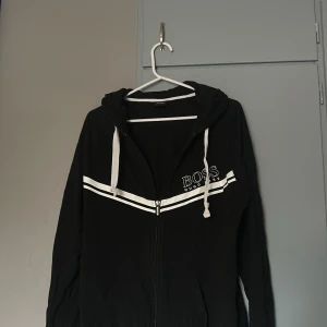 Svart hoodie från Hugo Boss - Svart hoodie från Hugo Boss med vita snören och två vita ränder över bröstet. Dragkedja framtill och huva. Boss-logga på vänster sida. Perfekt för en chill och stilren look.