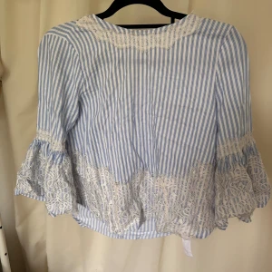 Blårandig blus med spets och rosett - Supersöt blus med blå och vita ränder, volangärmar och vacker vit spetsdetalj längs ärmar och nederkant. Baksidan har en snygg v-ringning med stor rosett. Tillverkad i bomull och viskos för en luftig känsla. Perfekt för dig som gillar romantisk och trendig stil.