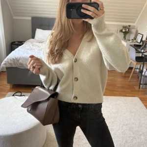 Beige kofta från Uniqlo - Supermjuk och mysig beige kofta från Uniqlo i storlek M. Koftan har v-ringning, tre knappar framtill och är lite croppad i modellen. Tillverkad i en stickad blandning med ull och polyester, perfekt för lager-på-lager under kyliga dagar.