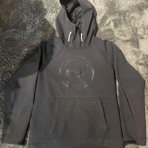 Svart skid hoodie från 8848 Altitude - Svart skid hoodie från 8848 Altitude med stor logga framtill. Tröjan har huva med vita snören, lång ärm och en stor magficka samt en inbyggd scarf som skyddar munnen från kylan. Materialet är mjukt och känns som syntet, perfekt för kyligare dagar. Dragkedja i sidan för enkel av- och påtagning.