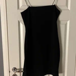 Svart kort klänning med kedjeband H&M - Säljer en svart kort klänning från H&M med smala kedjeband som axelband. Klänningen är ärmlös och har en enkel, stilren design som sitter snyggt på kroppen. Perfekt för dig som gillar minimalistisk stil med en liten edge.